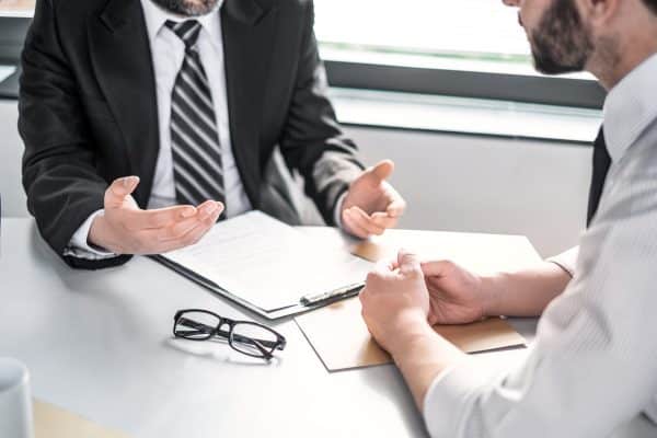 A lawyer talking to a client, with a clipboard and pair of glasses on the desk between them.