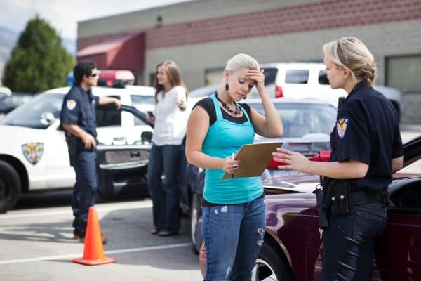 A distressed woman holding a clipboard and pen while talking to police after a car accident. Another woman speaks to another officer in the background.