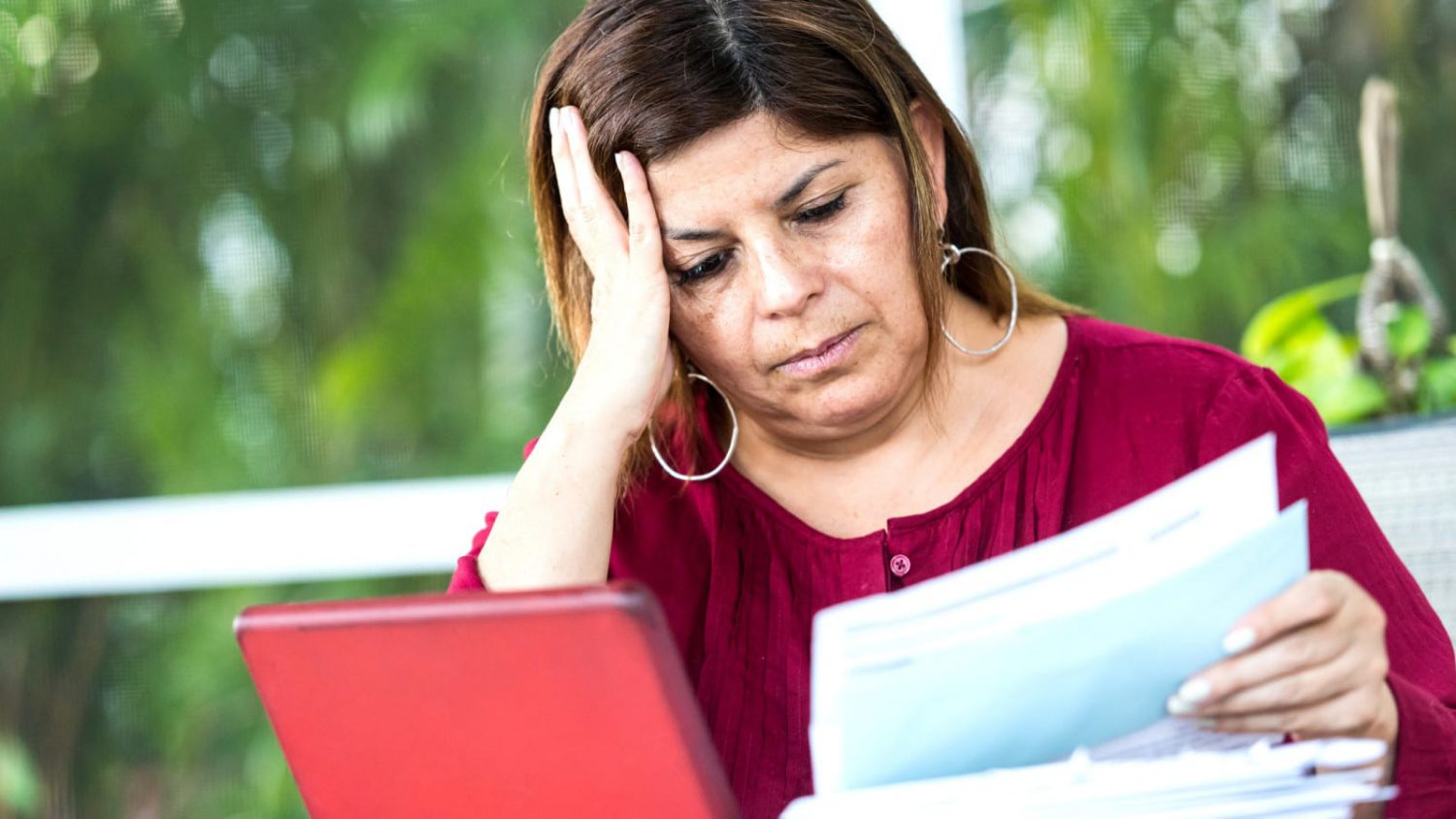 A distressed woman sits in front of a window with her hand on her face, reading some documents.