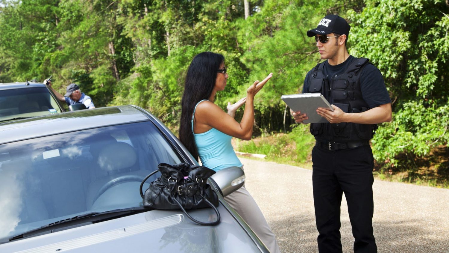 A police officer speaks with a woman after a car accident while the other driver is seen in the background examining his vehicle.