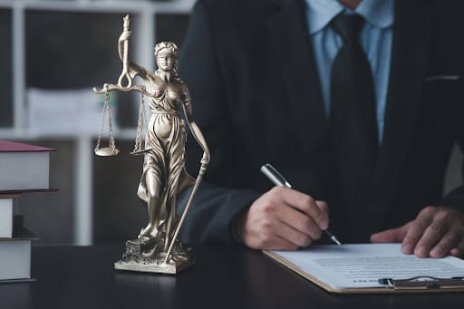An attorney sits at their desk, filling out a document on a clipboard. A statuette of Lady Justice is on the desk next to his paperwork.