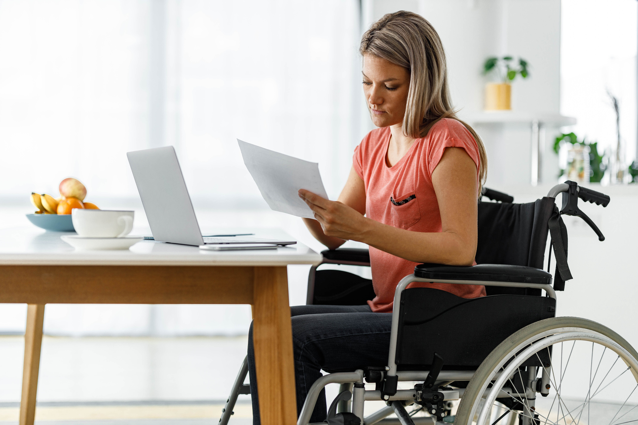 A woman in a wheelchair sits at a table in front of her laptop reviewing documents.